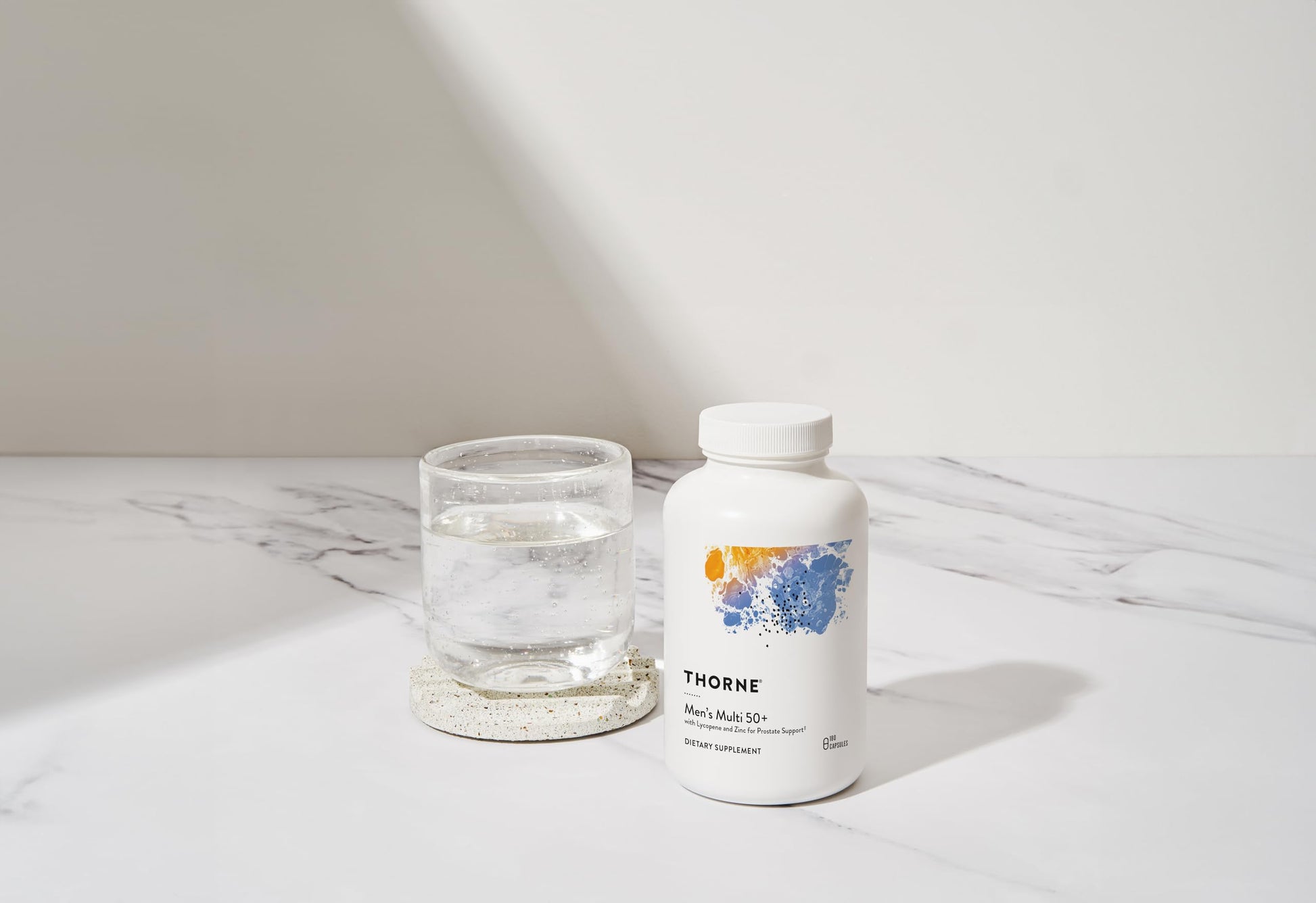 Botella de multivitamínico THORNE en escena minimalista junto a un vaso de agua.